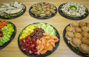 Six party platters on a wood table: sandwiches, croissant sandwiches, finger sandwiches, vegetables, fruit, sliders.