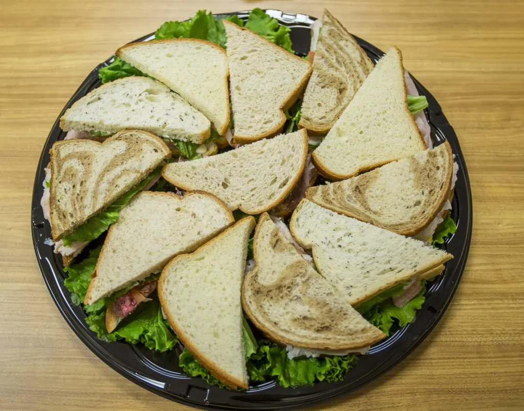 Overhead view of a round platter of sandwich halves on white and marble rye bread over lettuce on a wood table.
