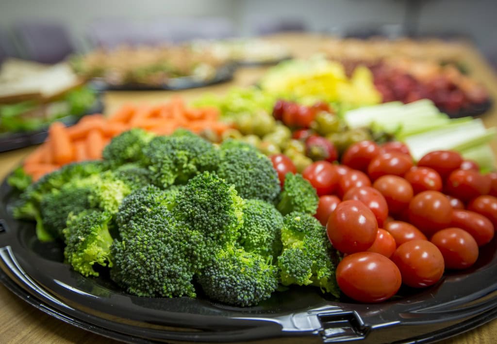 Vegetable tray with broccoli florets and cherry tomatoes in focus, carrots, olives and other platters behind.