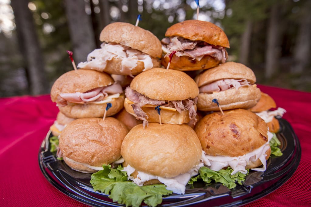 Tray of turkey, ham and roast beef sliders stacked in a pyramid on a red tablecloth with trees behind.