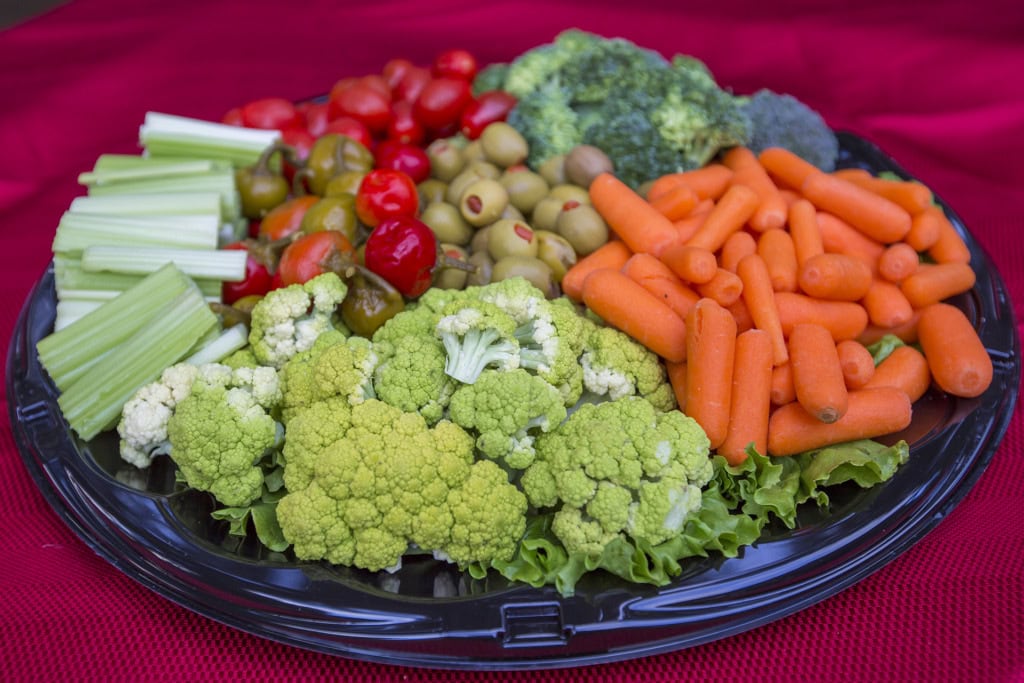 Vegetable tray with celery, cherry tomatoes, olives, baby carrots, broccoli and green cauliflower on a red cloth.