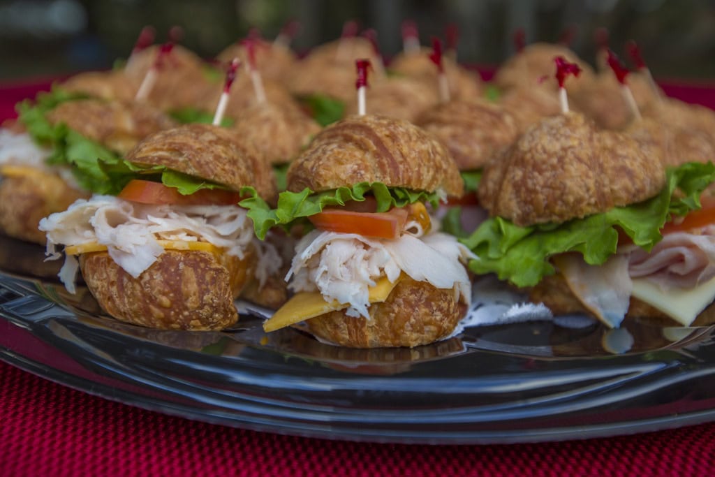 Close-up of mini croissant sandwiches with shaved turkey, cheese, tomato and lettuce on a black tray.