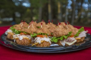 Tray of mini croissant sandwiches with turkey, ham, tomato and lettuce on a red cloth, trees blurred behind.