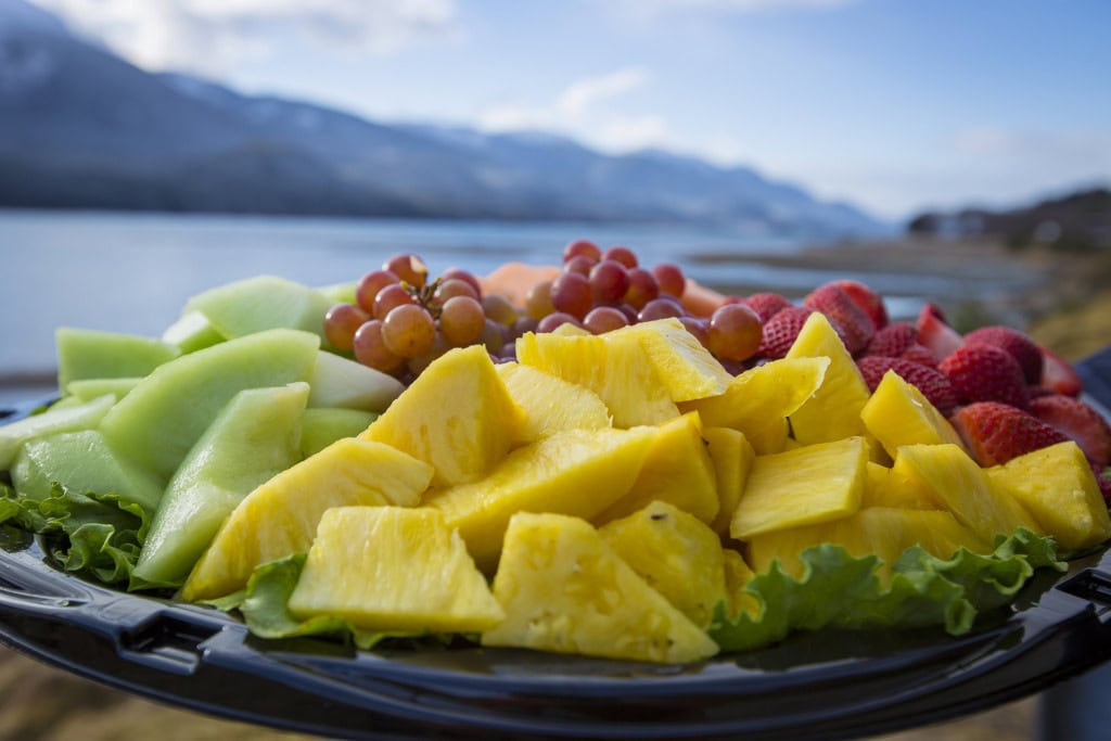 Fruit platter of pineapple chunks, honeydew, red grapes and strawberries, mountains and water out of focus behind.
