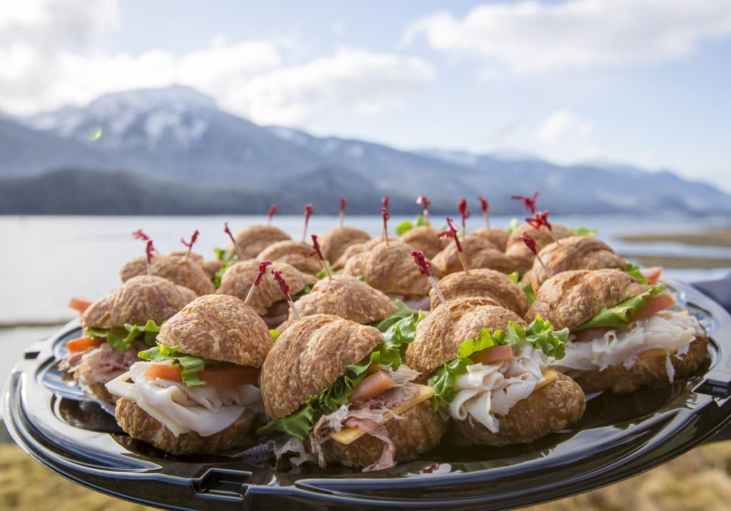 Platter of mini croissant sandwiches with turkey, ham, tomato and lettuce, snowy mountains and water behind.