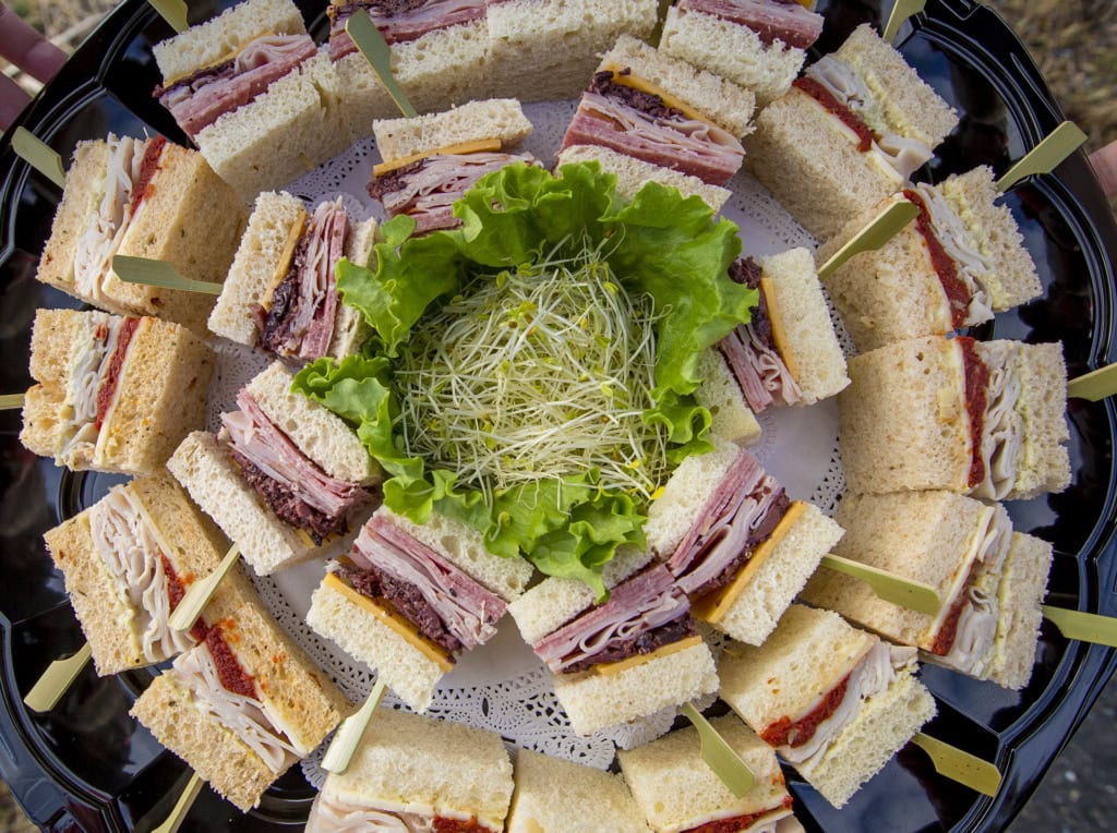 Overhead view of a round tray of cut finger sandwiches with turkey and ham, sprouts and lettuce in the center.