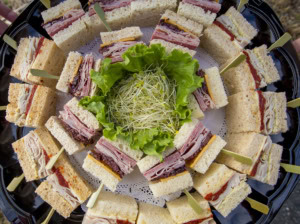 Overhead view of a round tray of cut finger sandwiches with turkey and ham, sprouts and lettuce in the center.