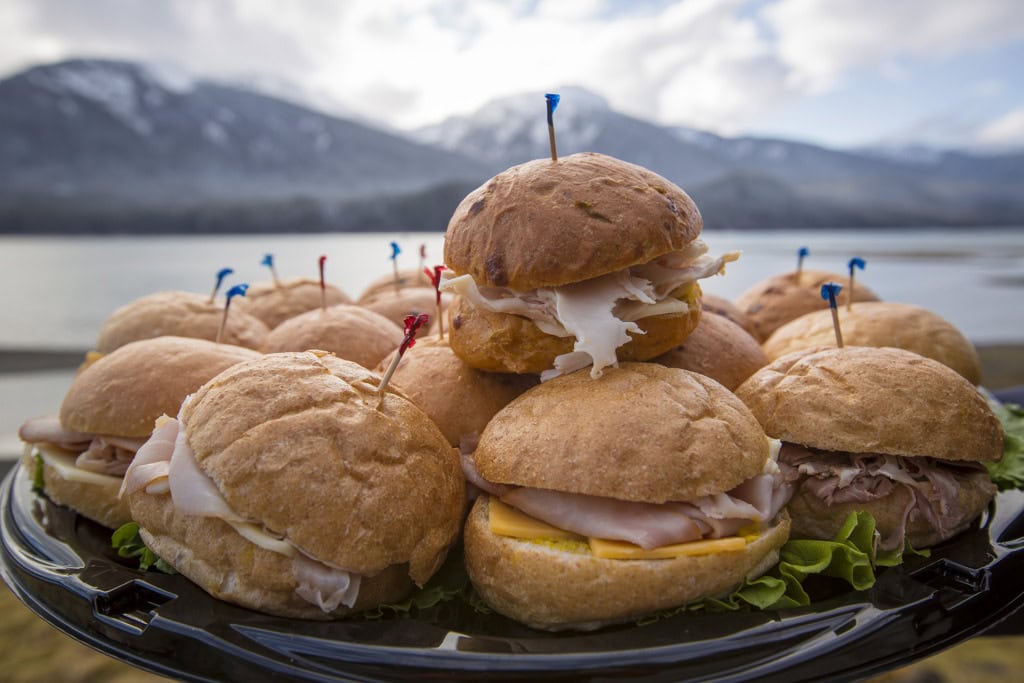 Platter of turkey and ham slider sandwiches stacked high, snow-capped mountains and calm water in the background.