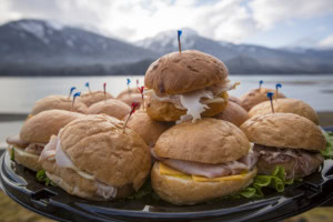 Platter of turkey and ham slider sandwiches stacked high, snow-capped mountains and calm water in the background.