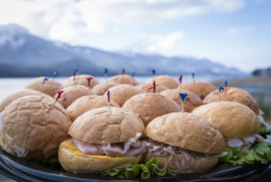 Tray of sliders with ham, roast beef and cheese on soft rolls, colored toothpicks, mountains and water behind.