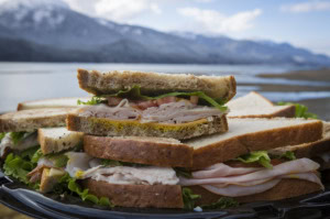 Turkey, cheese, lettuce and tomato sandwich halves stacked on a platter with snowy mountains and water behind.