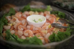 Platter of peeled shrimp with lemon wedges and lettuce around a cup of creamy dip, tongs resting on the side.