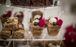 Mini dessert cups topped with cream, a raspberry and a blueberry next to stacked blondie bars on an acrylic stand.