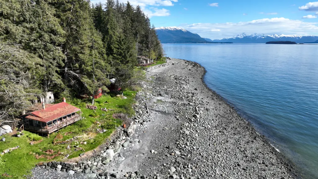 Aerial view of a red-roofed cabin on a forested point beside a long gravel spit and calm channel water