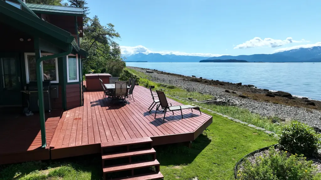 A red cedar deck and lawn overlooking a rocky beach, calm water and distant mountains
