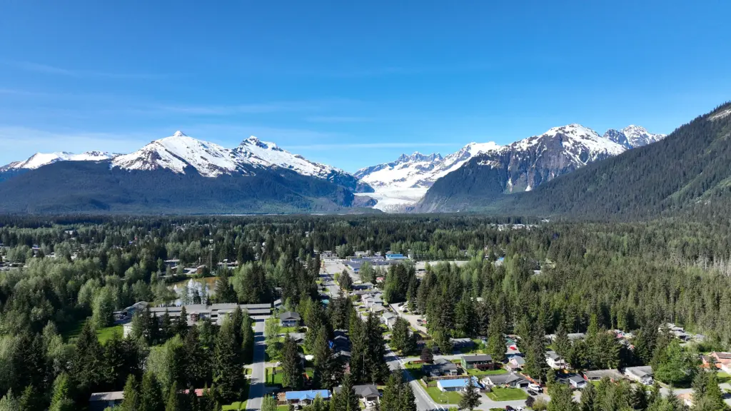 Aerial view over a tree-lined Mendenhall Valley neighborhood toward Mendenhall Glacier and snowy peaks