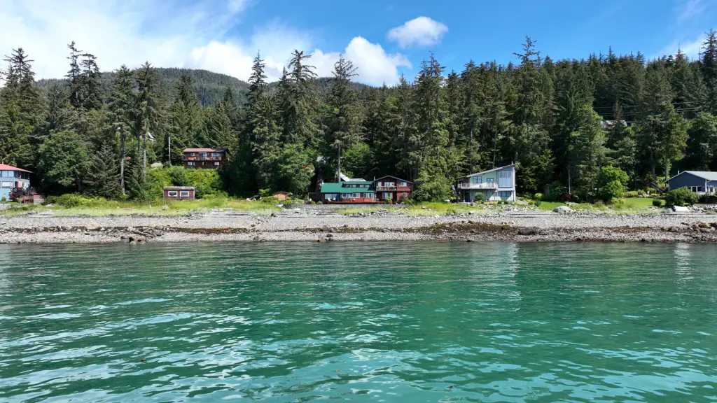 Clear turquoise water in front of a rocky beach and a row of homes beneath tall spruce