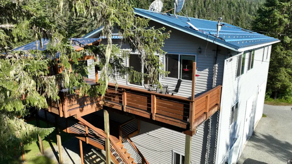 A wide wooden deck and staircase wrapping a home surrounded by spruce trees