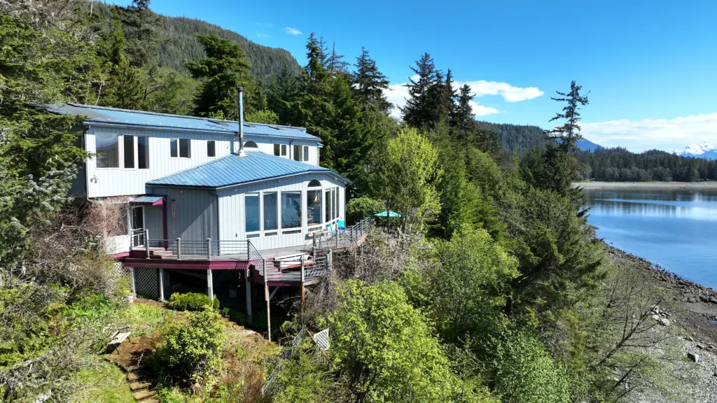 A white home perched on a forested bluff above a gravel beach and calm inlet