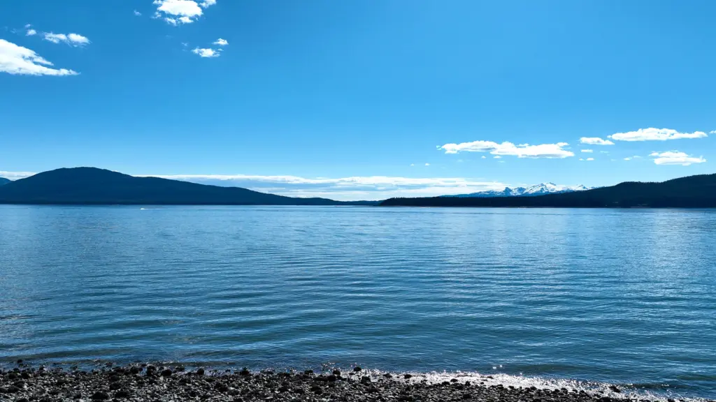 Calm blue channel water with forested islands and distant snowcapped peaks, seen from a pebble beach