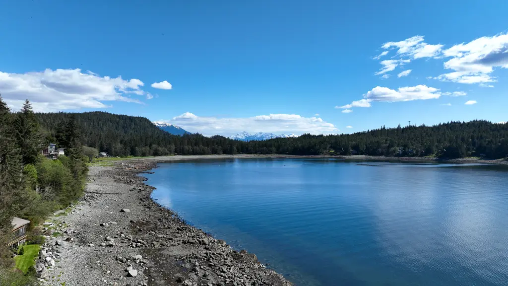 A curving gravel beach and calm blue cove framed by forest and distant mountains