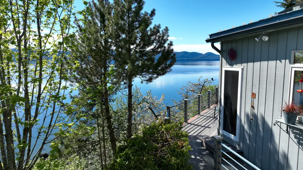 A home's deck and siding beside tall trees with a wide view of blue water and islands