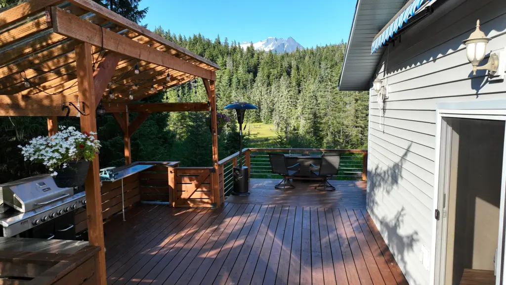 A wooden deck with pergola, grill and seating beside a home with forest and mountains behind