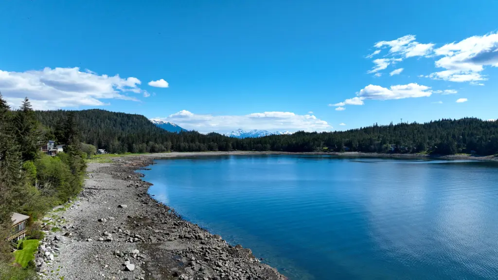 A curving rocky beach and calm blue cove ringed by forest and waterfront homes, with snowy peaks beyond