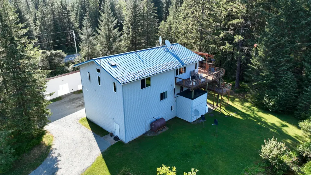 A white two-story home with a metal roof, deck, lawn and gravel drive among spruce trees