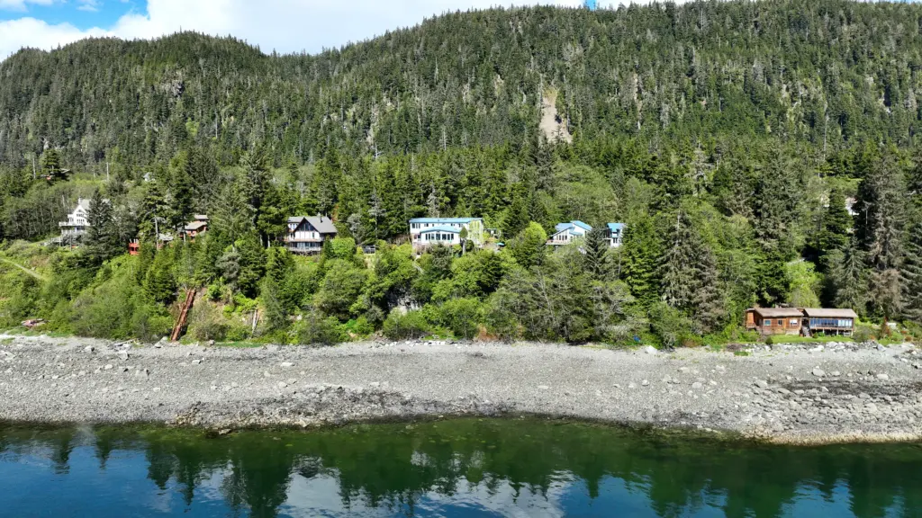 A row of homes tucked into a forested hillside above a gravel beach and still water