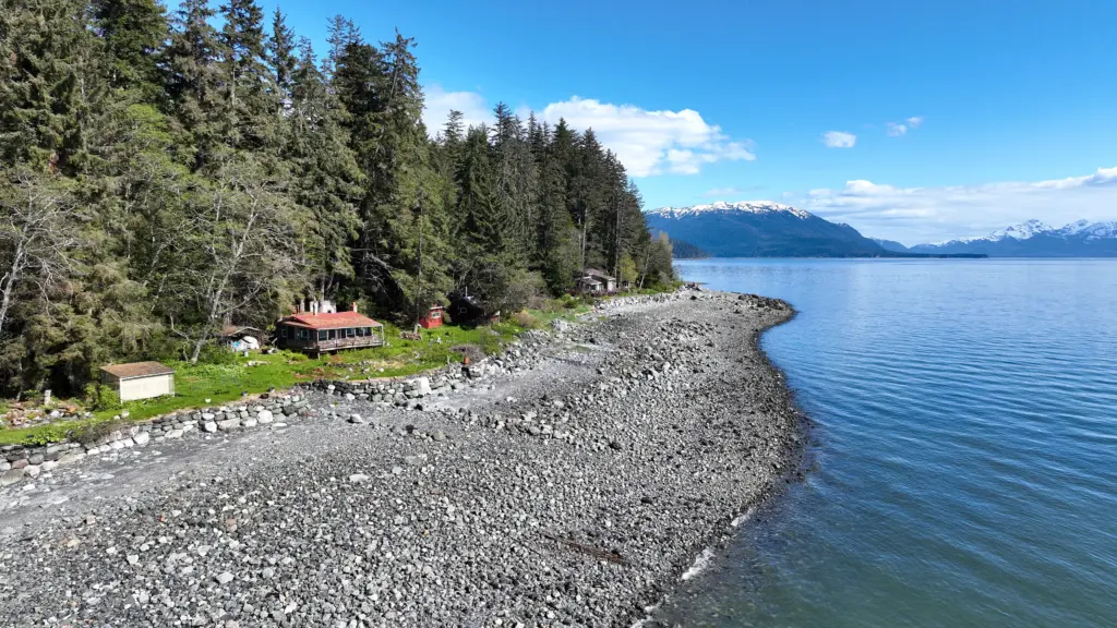 A long gravel spit curving into calm blue water beside a cabin at the forest edge