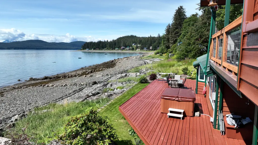 A red deck with a hot tub and chairs looking out over a rocky beach and calm water