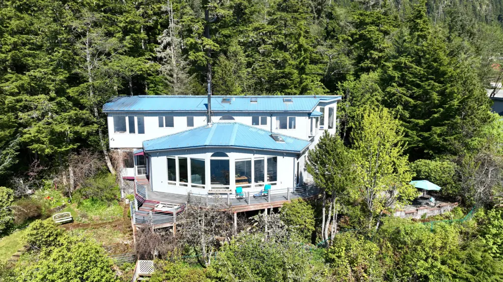A white home with a blue roof and large windows set among spring-green trees on a hillside