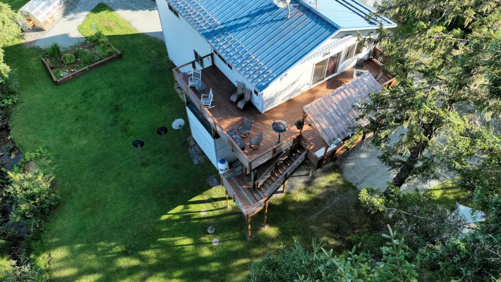 An aerial view of a home's metal roof, decks and green lawn beside tall trees