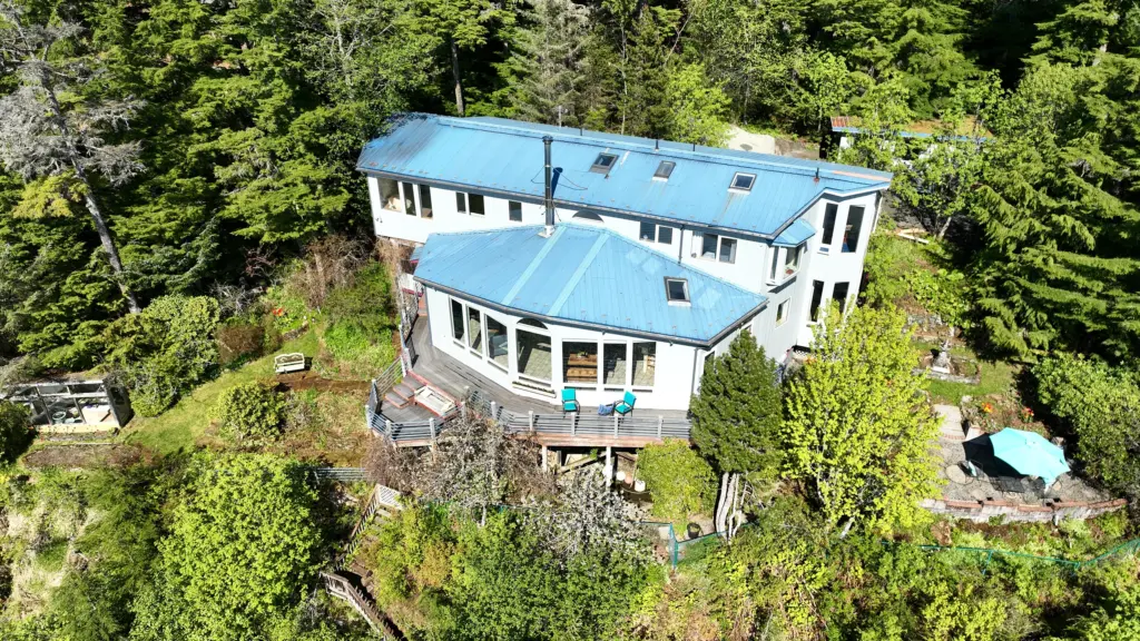 Aerial view of a blue-roofed hillside home with decks, gardens and forest